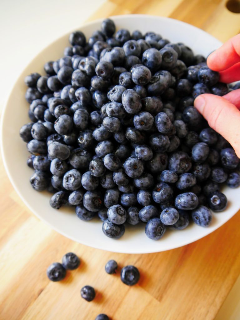 Blueberries fill a white bowl, which is set on a wooden board. In the upper right corner of the picture, a hand is just visible, snitching a few berries from the bowl.