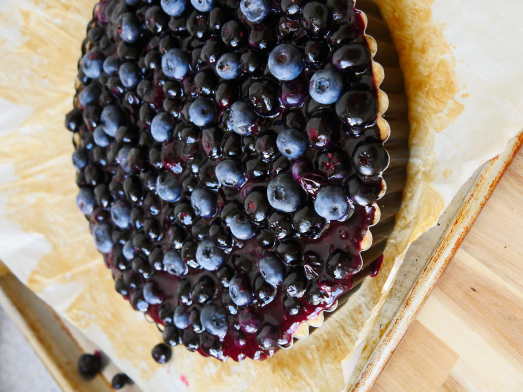 A glossy purple blueberry tart, topped with fresh raw berries, rests on a parchment paper-covered baking sheet.